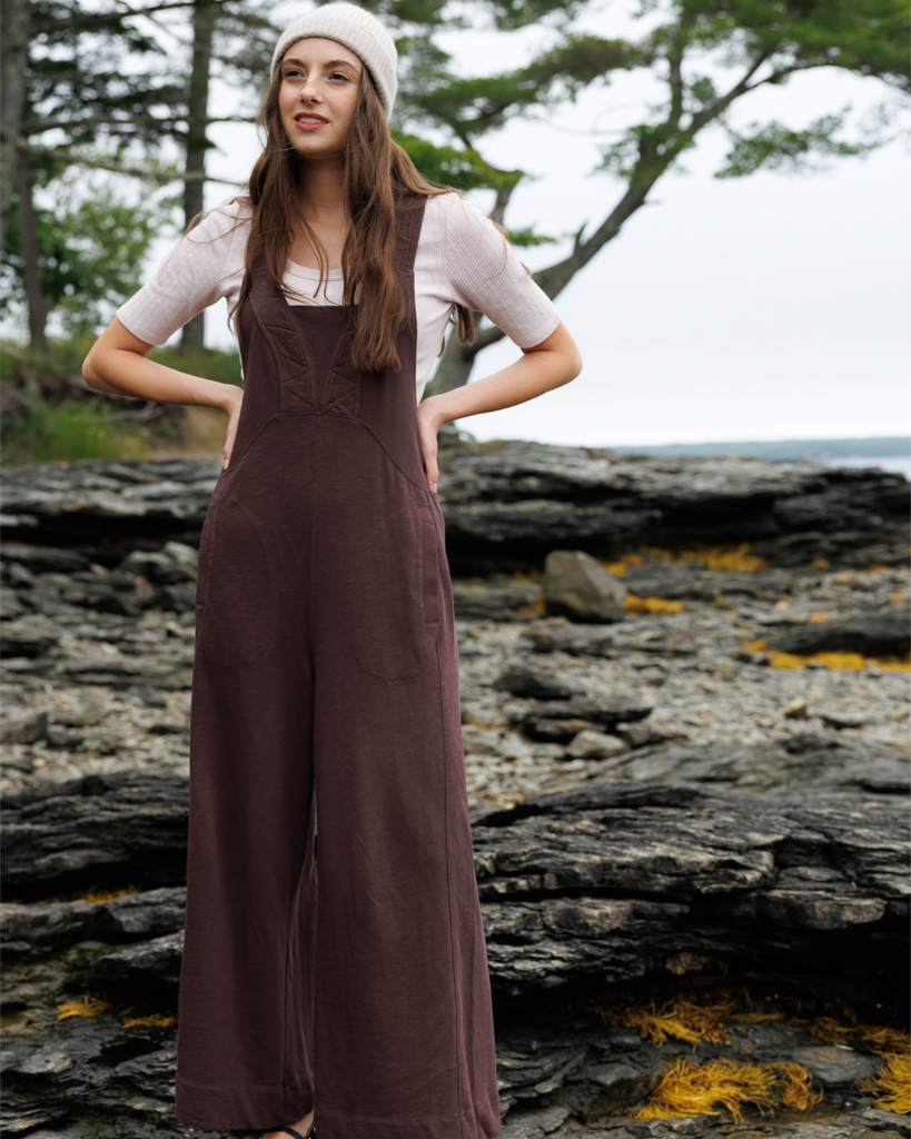 Woman wearing a brown jumpsuit and pink undershirt standing on rocks outside.