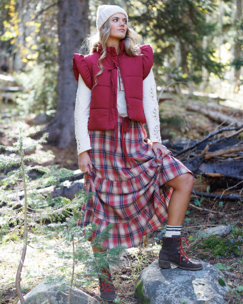 Woman in a red plaid skirt and puffer vest standing in a forest.