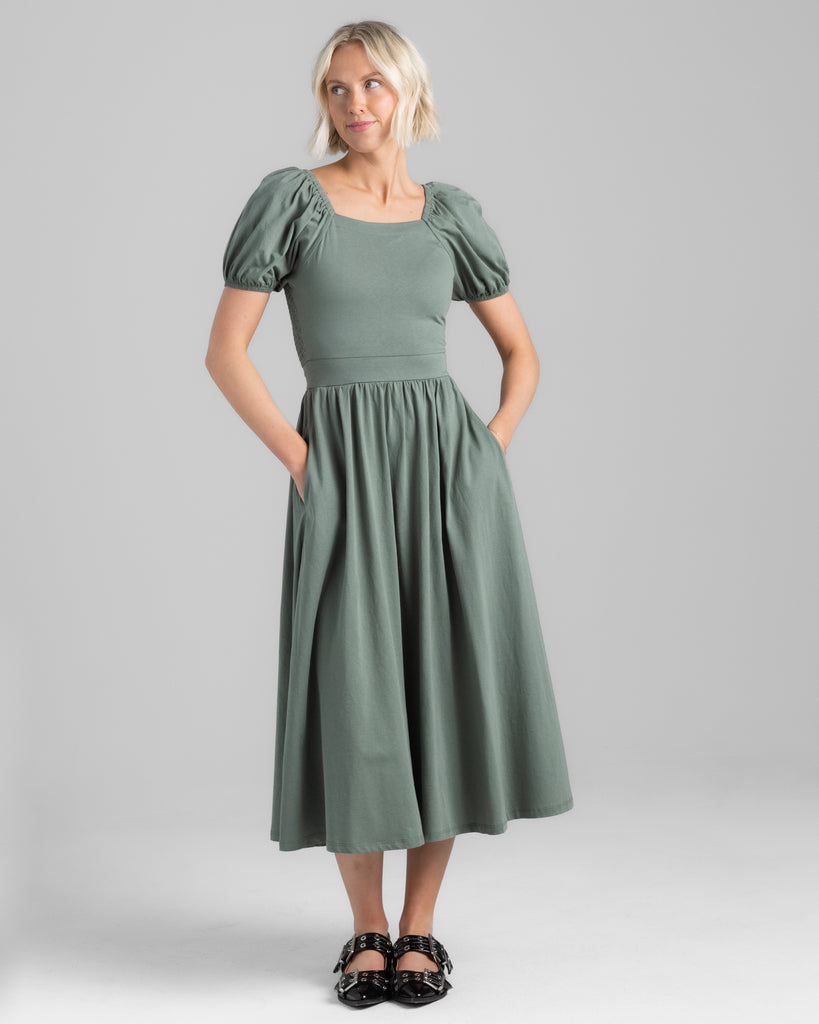 Model wearing a sage green, short puffed sleeve, square neck midi length dress with pockets.