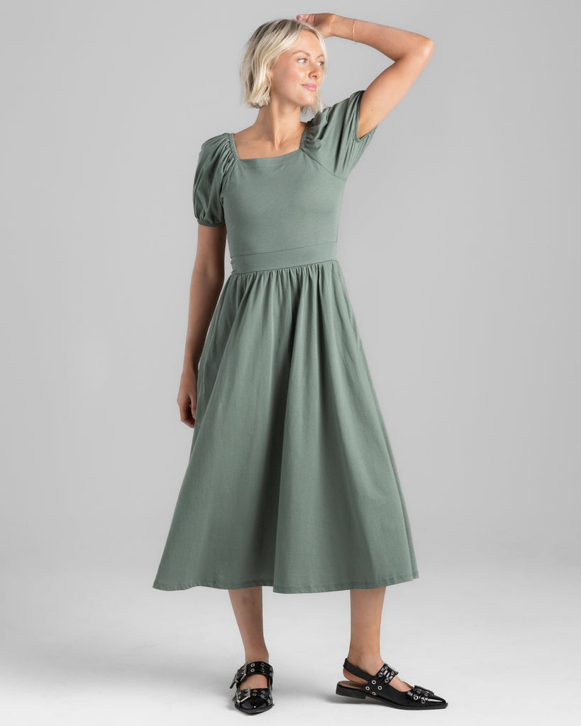 Model wearing a sage green, short puffed sleeve, square neck midi length dress with pockets.