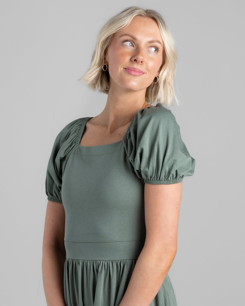 Model wearing a sage green, short puffed sleeve, square neck midi length dress with pockets.