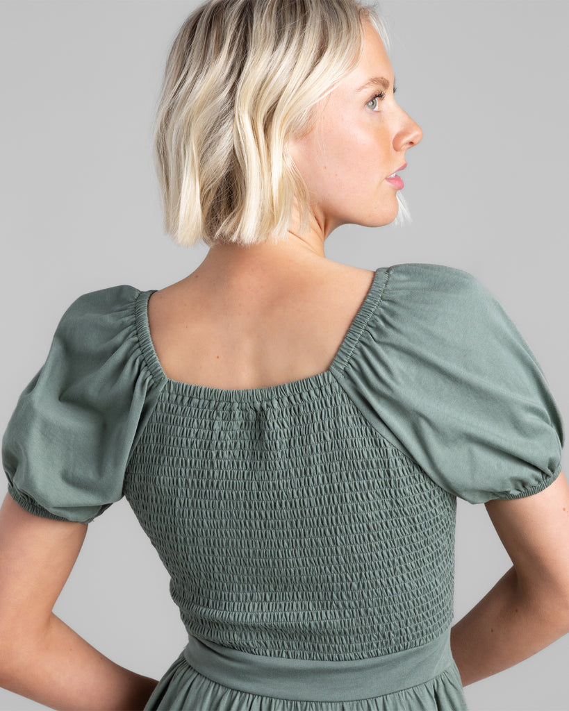 Model wearing a sage green, short puffed sleeve dress with smocking on back of bodice.