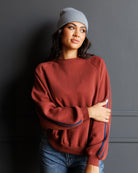 A woman wearing a rust-colored crewneck sweatshirt with blue stripes on the sleeves, paired with a gray beanie and blue jeans, posing against a dark gray wall.