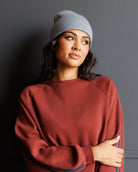 A woman wearing a rust-colored crewneck sweatshirt with blue stripes on the sleeves, paired with a gray beanie and blue jeans, posing against a dark gray wall.