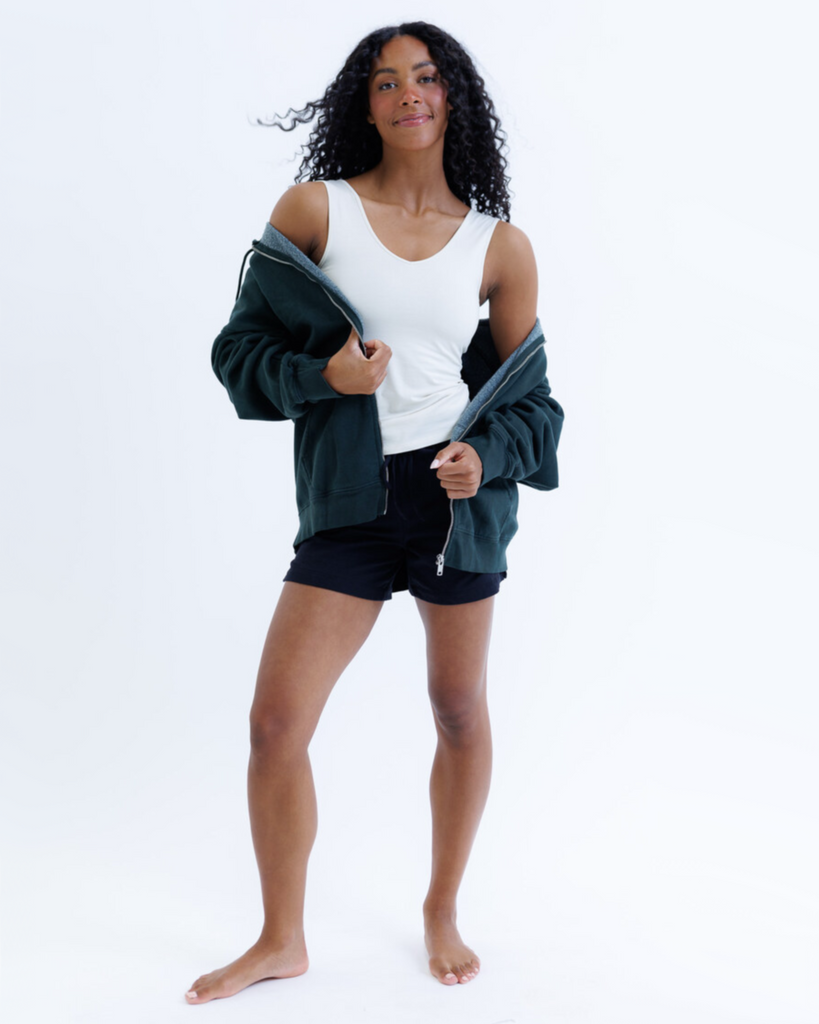 Woman wearing a cream colored layering tank top and black shorts with a dark jacket on a white background