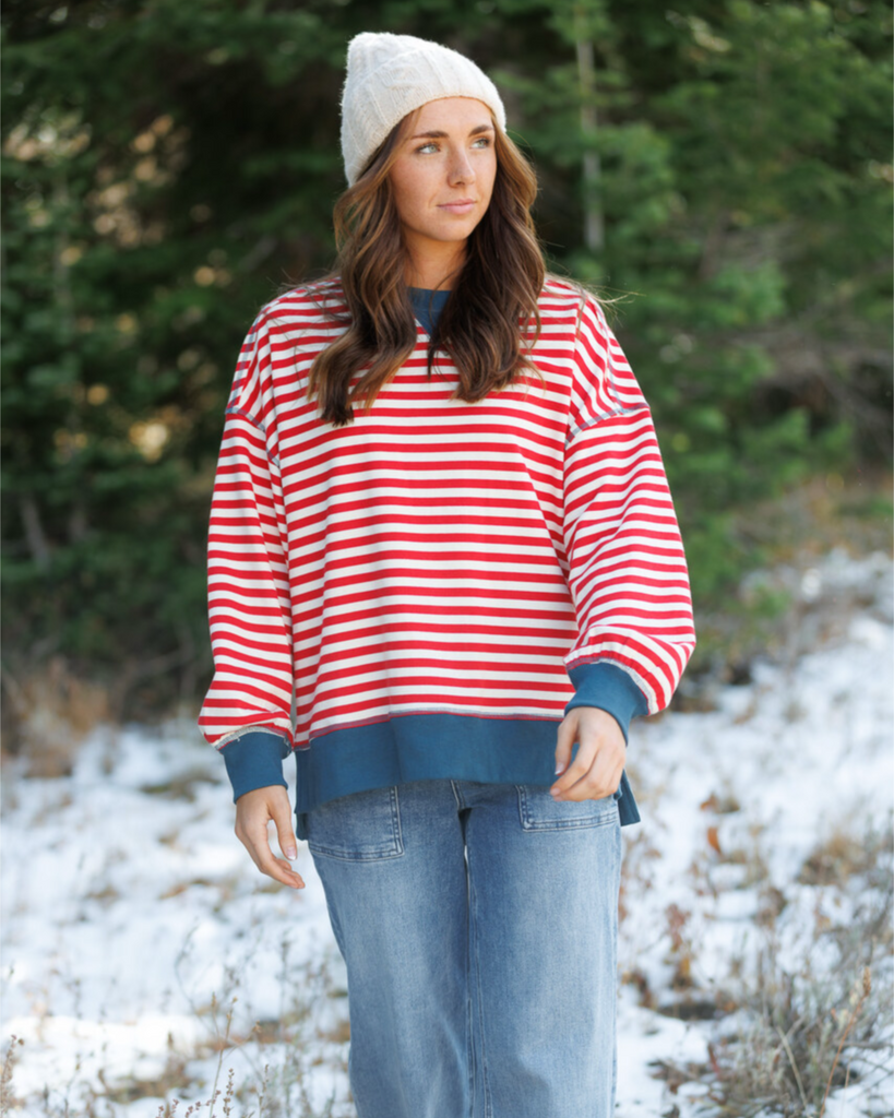 Woman standing in forest wearing a beanie, red and blue striped sweatshirt, and light denim jeans
