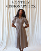 A woman stands against a draped cream fabric backdrop wearing a short-sleeve button-front midi dress in a light taupe-beige color. Large text at the top reads 'MONTHLY MISSIONARY BOX'.