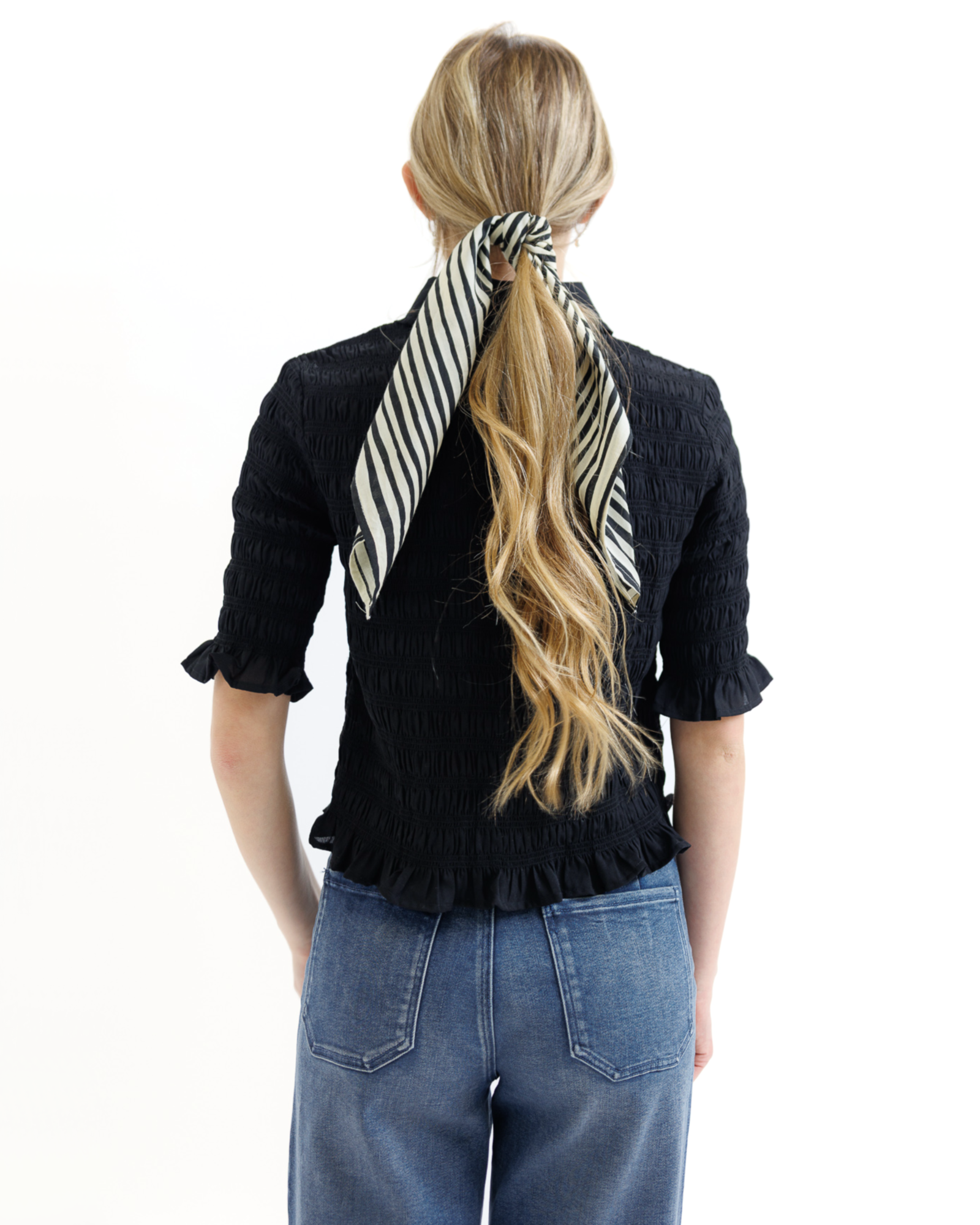 Woman wearing a black textured blouse and blue jeans with a striped hair accessory on a white background