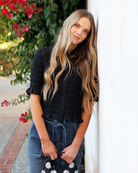 Woman in a black textured blouse and blue jeans leaning against a white wall with flowers in the background