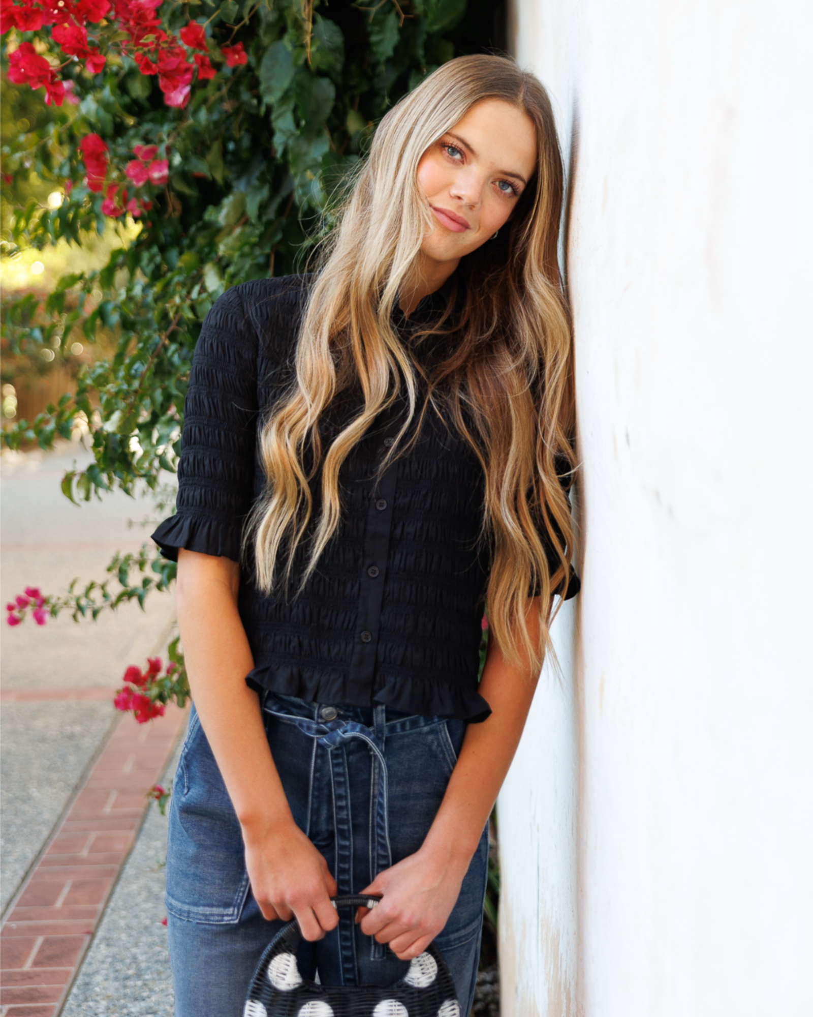 Woman in a black textured blouse and blue jeans leaning against a white wall with flowers in the background