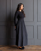 A woman modeling a long-sleeve black maxi dress with a pleated skirt, standing against a dark gray paneled wall on a wooden floor.