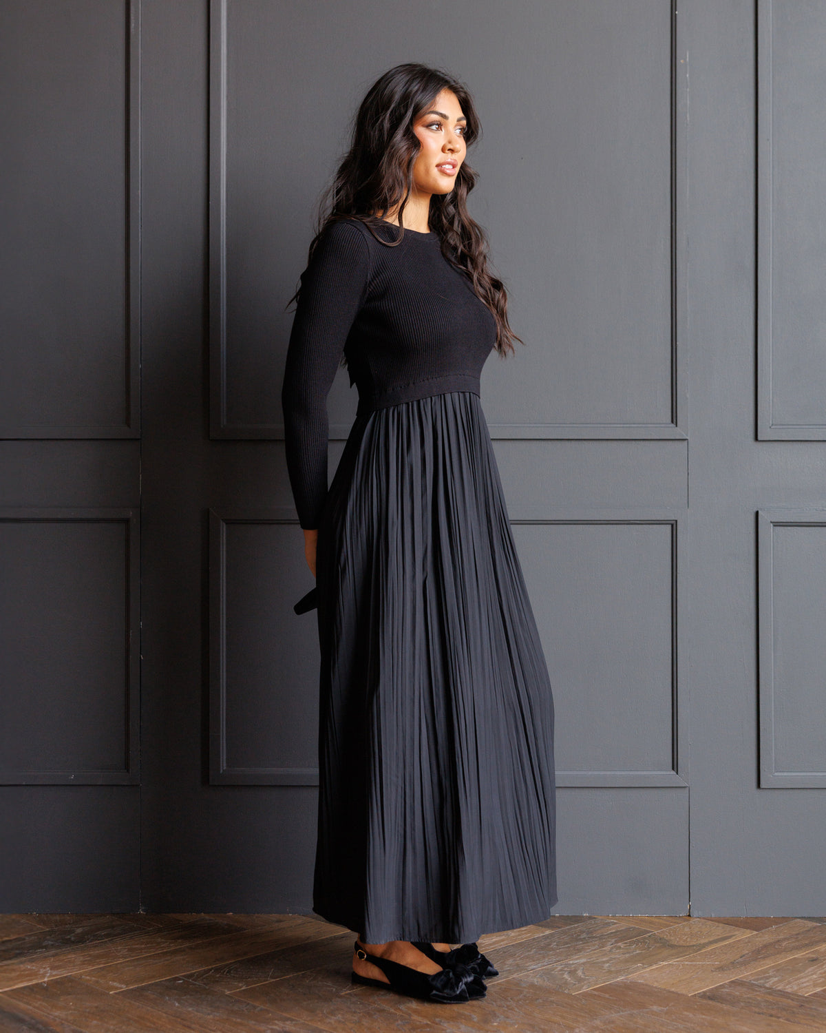 A woman modeling a long-sleeve black maxi dress with a pleated skirt, standing against a dark gray paneled wall on a wooden floor.