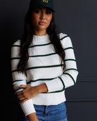 A woman wearing a white ribbed knit sweater with dark green horizontal stripes, paired with blue jeans and a dark green baseball cap, posed against a dark gray wall.