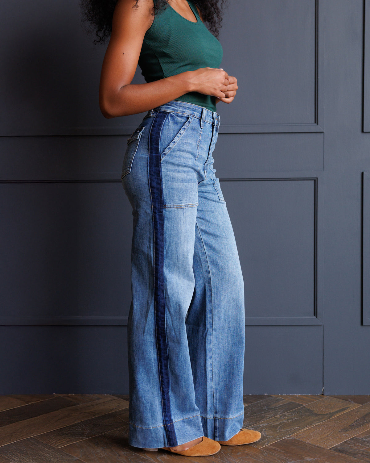 A model wearing high-waisted, wide-leg blue denim jeans with dark blue vertical stripe detailing down the sides, paired with a green tank top and tan shoes, standing against a dark gray paneled wall.