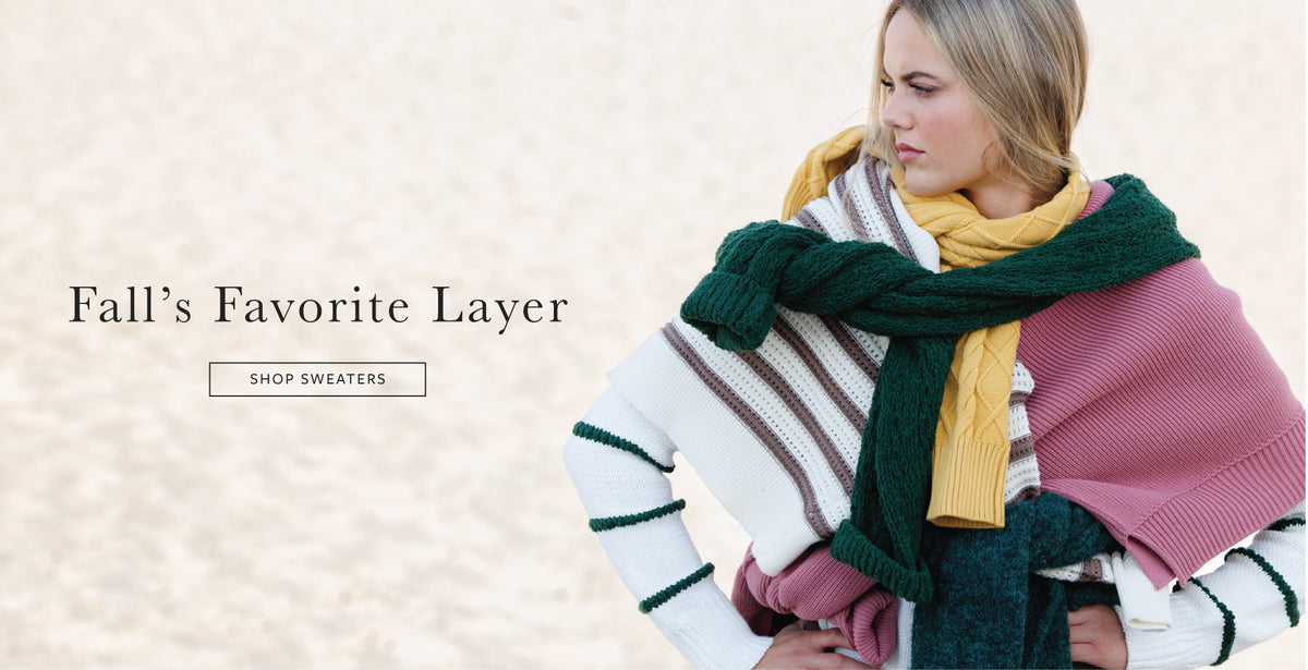 A woman wearing a layered knit sweater with a striped white, brown, and green body, styled with a mustard yellow scarf and a dark green knit wrap, against a light beige background with the text 'Fall's Favorite Layer' and 'SHOP SWEATERS'.