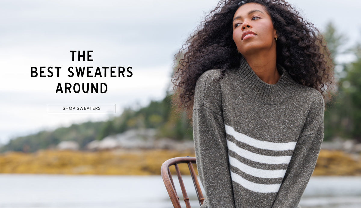 A woman wearing a gray knit sweater with three white horizontal stripes on the front, seated on a wooden chair outdoors near water and trees. Text on the left reads 'THE BEST SWEATERS AROUND' with a 'SHOP SWEATERS' button below.