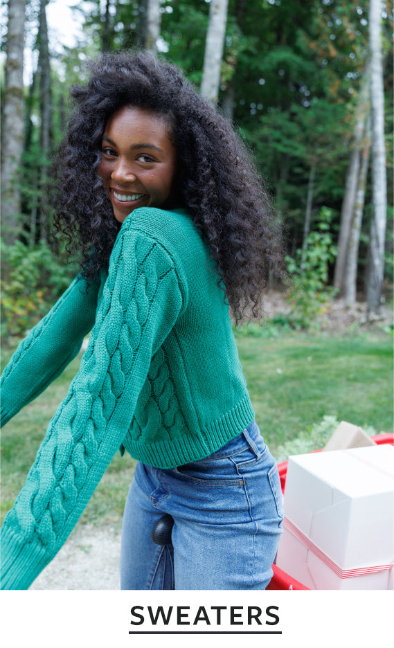 A woman outdoors wearing a green cable-knit cropped sweater and blue jeans, with trees and grass in the background. The word 'SWEATERS' appears in a black banner at the bottom.