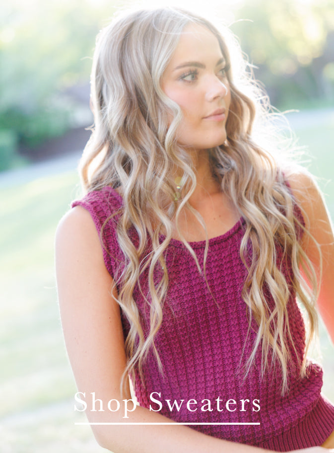 A woman with long, wavy blonde hair wearing a sleeveless, textured magenta knit sweater, set against a blurred outdoor background with a text overlay at the bottom reading 'Shop Sweaters'.