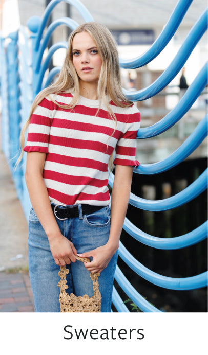 Woman wearing a red and white striped sweater with blue jeans, standing against a blue architectural background.