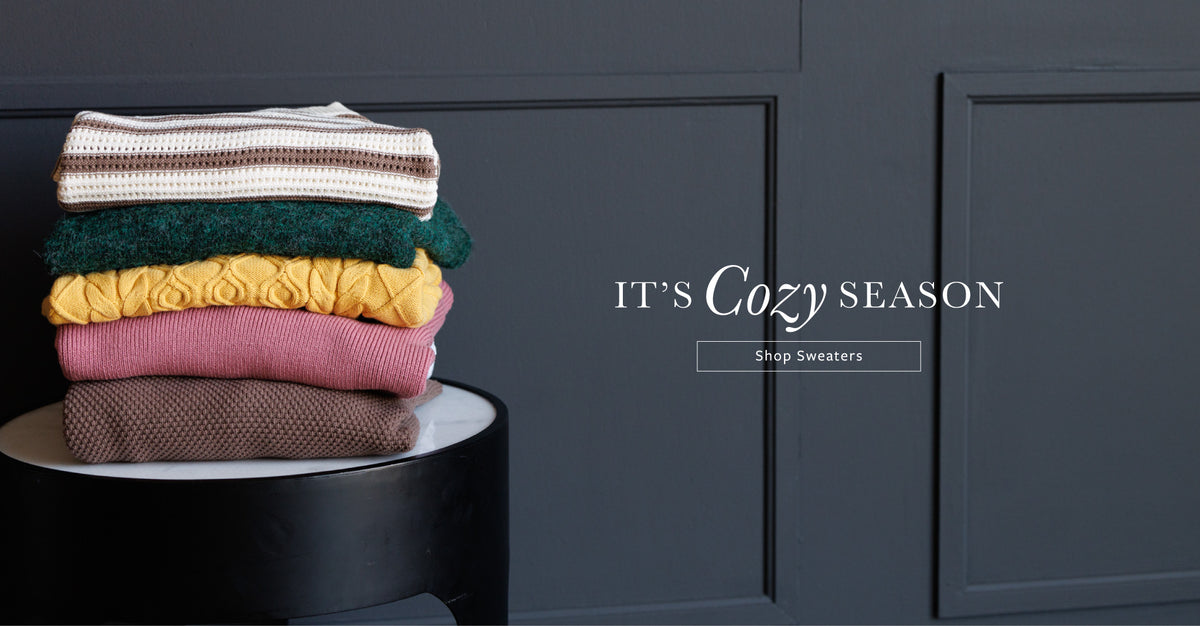 A stack of five folded knit sweaters in cream, green, yellow, pink, and brown sits on a black side table against a dark gray paneled wall. White text on the right reads 'IT'S Cozy SEASON' with a smaller 'Shop Sweaters' button.