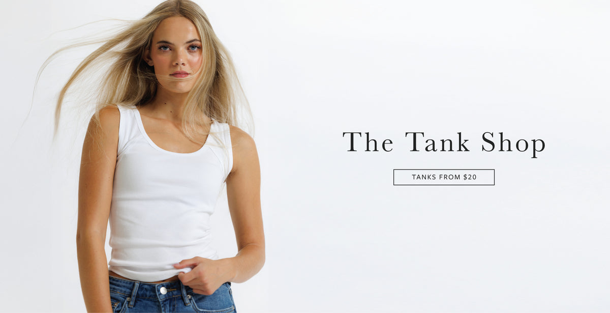 A woman modeling a white tank top against a plain white background, with the text 'The Tank Shop' and 'TANKS FROM $20' printed on the right side.