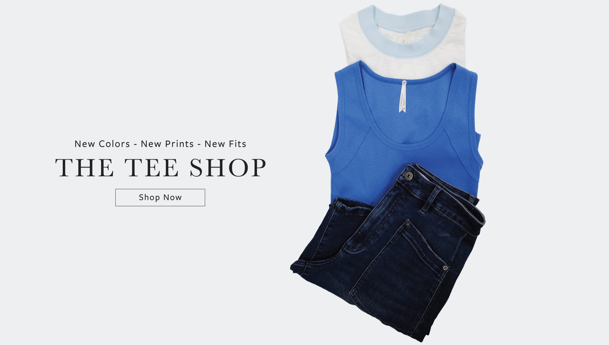A styled flat-lay of folded clothing on a light gray background, featuring a bright blue sleeveless top layered over a white crewneck tee and dark blue jeans. Text on the left reads 'New Colors - New Prints - New Fits THE TEE SHOP' with a 'Shop Now' button.