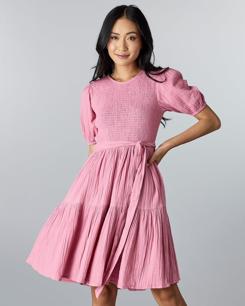 Woman in a pink, short sleeved, knee-length dress with smocking on bodice and a tie at the waist.