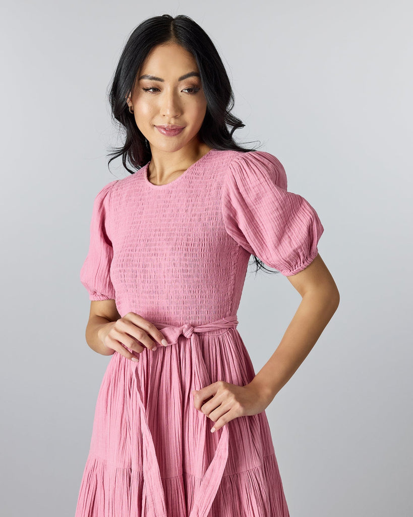 Woman in a pink, short sleeved, knee-length dress with smocking on bodice and a tie at the waist.