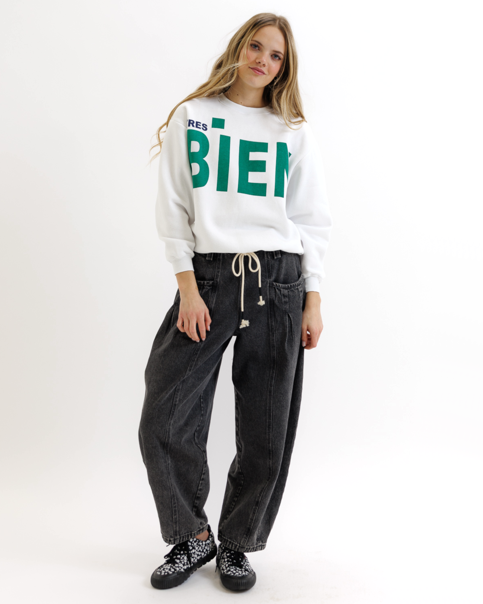 Woman wearing a white sweatshirt with green text that says 'TRES BIEN' and black jeans on a white background