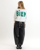 Woman wearing a white sweatshirt with green text that says 'TRES BIEN' and black pants on a white background