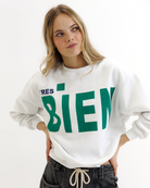 Woman wearing a white sweatshirt with green text that says 'TRES BIEN' on a white background