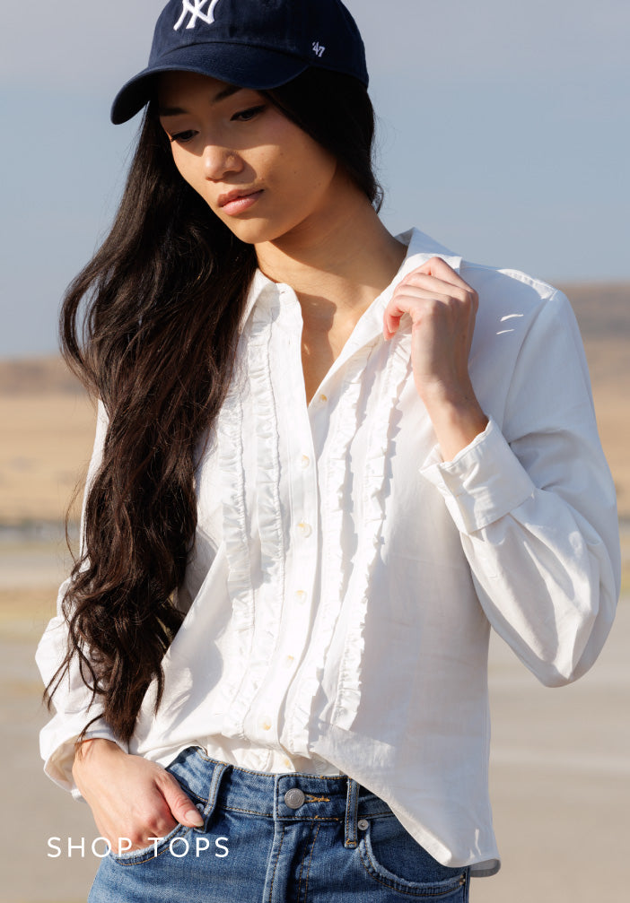 A woman wearing a white long-sleeve button-up blouse with ruffle trim, tucked into blue jeans, and wearing a navy baseball cap. The photo is taken outdoors in a desert setting.