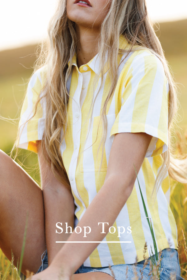 A woman wearing a yellow and white vertically striped short-sleeve button-up shirt, sitting outdoors in a grassy field. The text 'Shop Tops' is overlaid at the bottom.