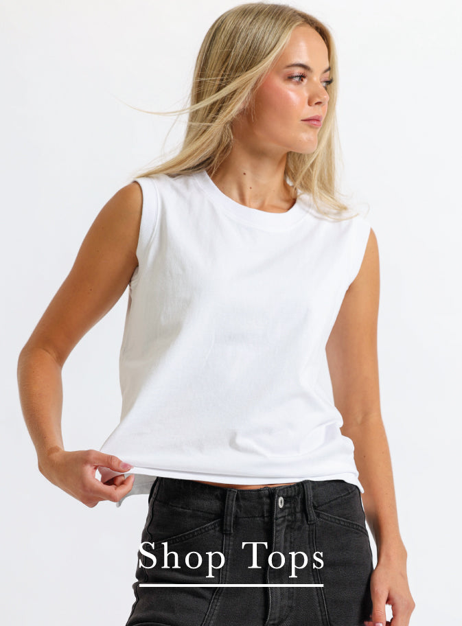 A blonde woman wearing a white sleeveless crewneck top and black jeans, posing against a white studio background with 'Shop Tops' text overlay at the bottom.