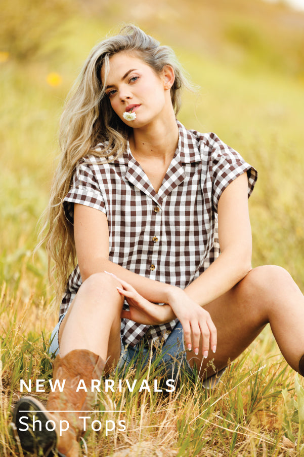 Woman sitting in a field wearing a checkered shirt with text overlay 'NEW ARRIVALS Shop Tops'.