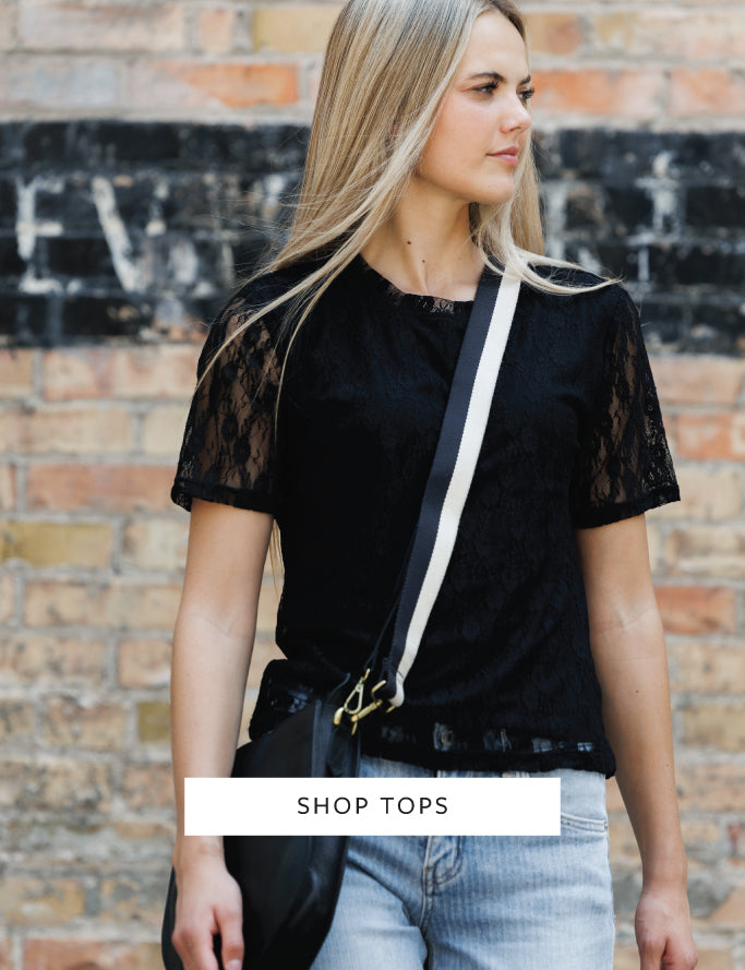 A woman wearing a black short-sleeve lace top and light blue jeans, carrying a black shoulder bag with a white strap, standing in front of a brick wall. A white banner at the bottom reads 'SHOP TOPS'.