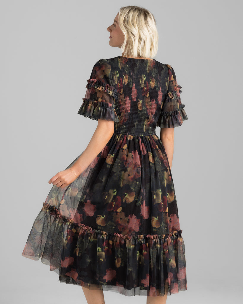 Model wearing a black with pink watercolor floral print, short sleeve with ruffles, v-neck, smocked bodice, midi length tulle dress.