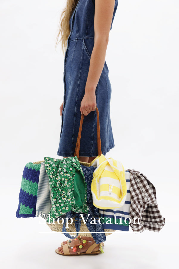 A person wearing a blue denim dress holds a large, colorful bundle of assorted travel accessories, including a striped knit blanket, a green floral tote bag, a yellow and white striped pillow, and a plaid shirt, against a plain white background.