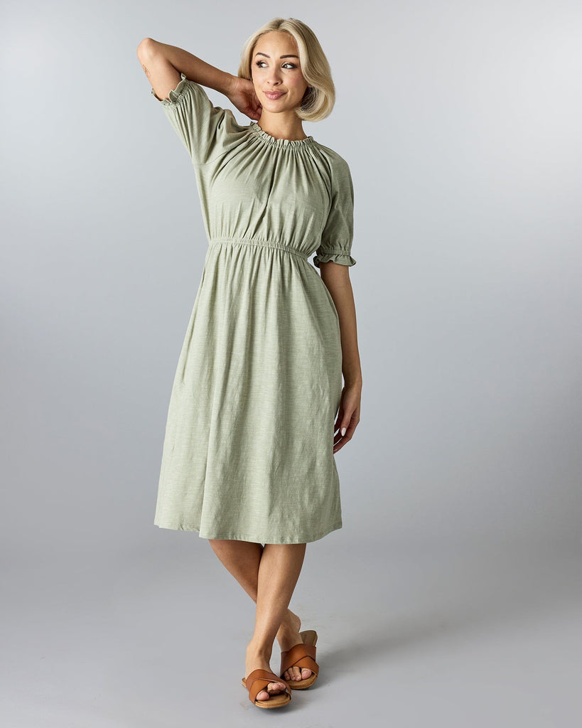 Woman in a green, knee-length, short sleeve dress with a high neckline and ruffles at neck and arms.