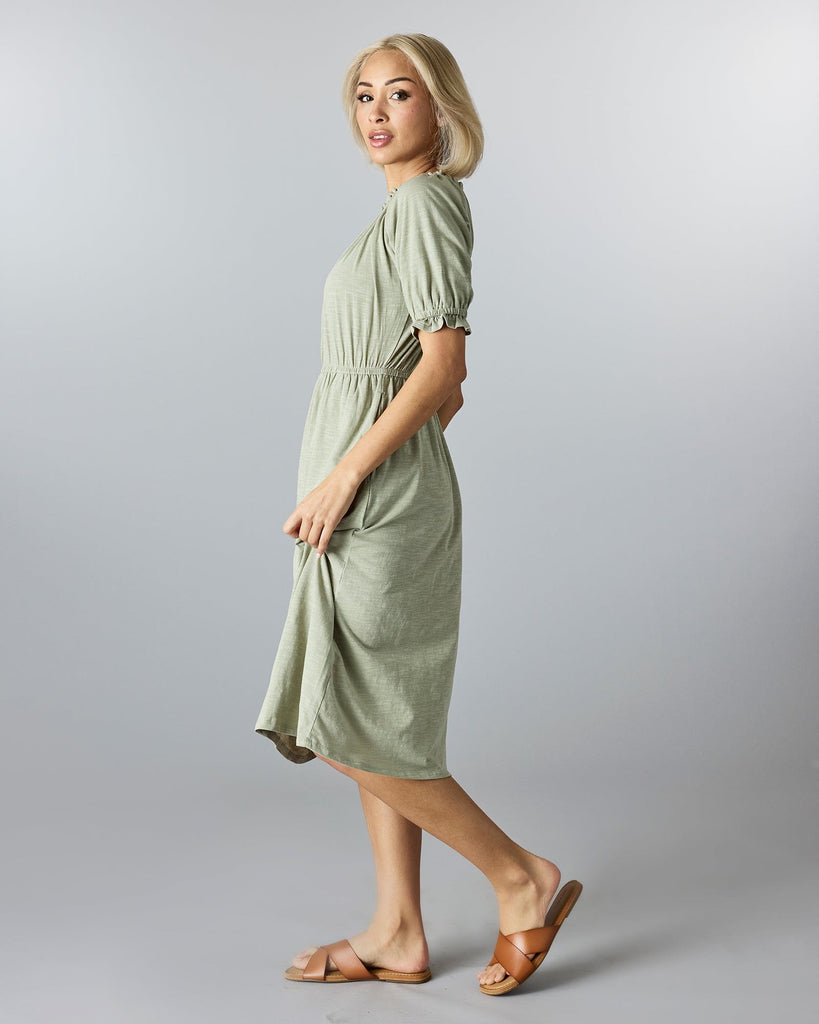 Woman in a green, knee-length, short sleeve dress with a high neckline and ruffles at neck and arms.