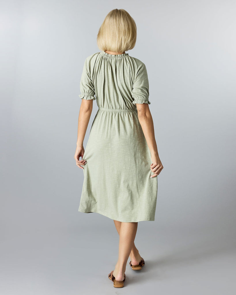 Woman in a green, knee-length, short sleeve dress with a high neckline and ruffles at neck and arms.