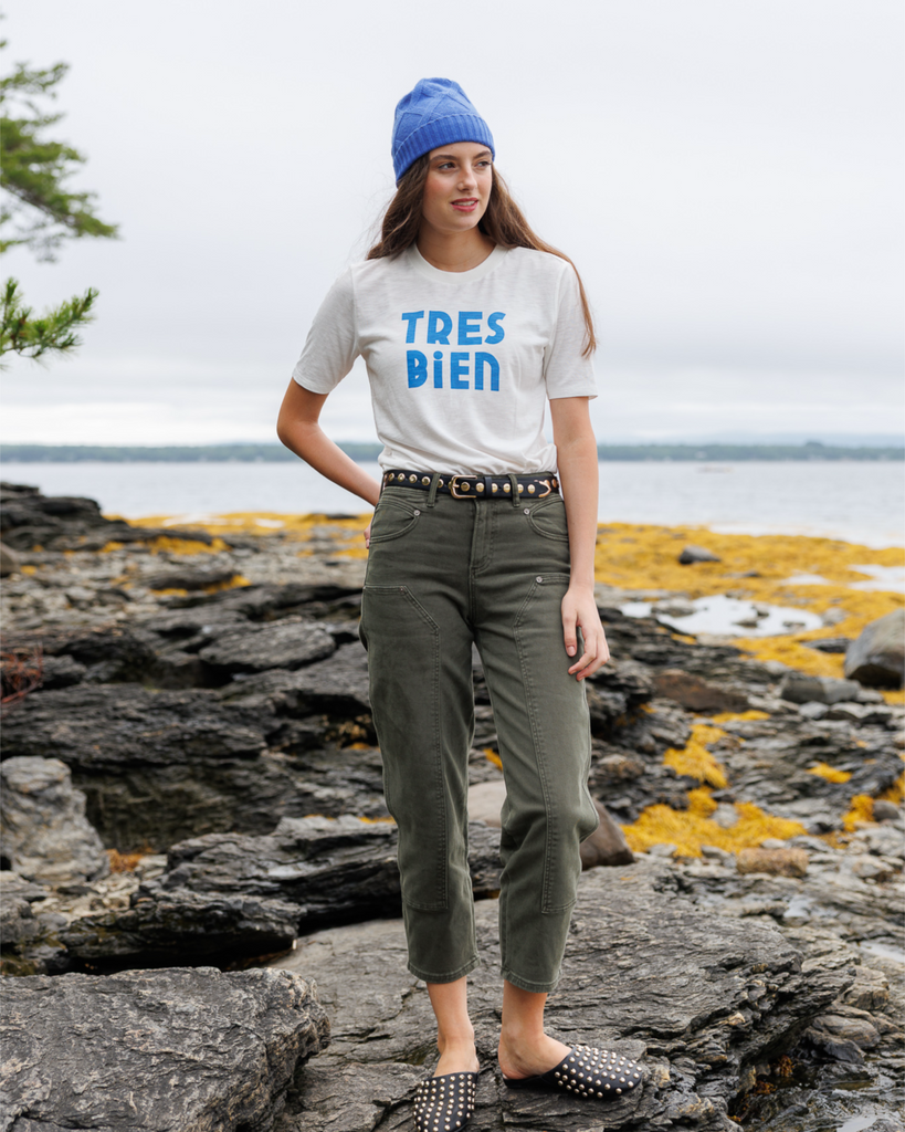 Woman wearing a blue beanie, white shirt, and green pants standing on rocks outside.