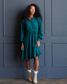 A woman with curly black hair wearing a teal green tiered midi dress with a tie waist and beige ankle boots, standing against a dark blue paneled wall.
