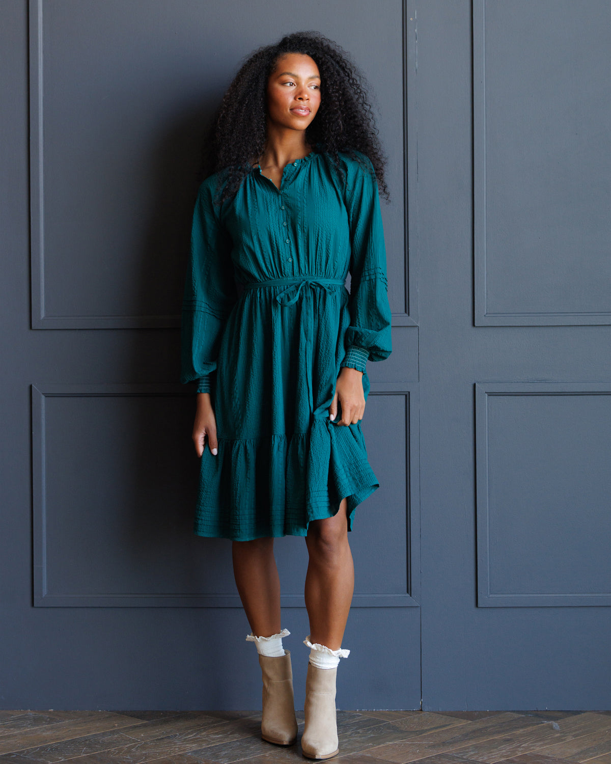 A woman with curly black hair wearing a teal green tiered midi dress with a tie waist and beige ankle boots, standing against a dark blue paneled wall.