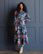 A woman with curly black hair models a long-sleeved navy blue midi dress with a colorful floral print, standing against a dark blue paneled wall.