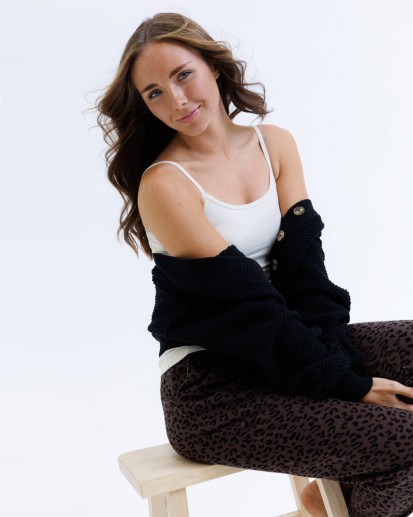 Woman wearing a black cardigan over a cream colored layering tank top and leopard print sweatpants on a white background