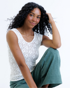Woman wearing a white cap sleeve top and green pants sitting on a white background