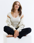 Woman sitting cross-legged on a white background wearing a cream colored layering top and black pants.