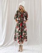 A woman stands against a white draped fabric backdrop wearing a dark floral midi dress with puff sleeves, a white knit beanie, white socks, and beige ankle boots.