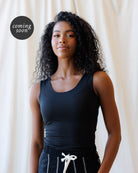A woman with curly dark hair wearing a black sleeveless scoop-neck top and black pants with a white drawstring, standing against a draped cream fabric backdrop. A circular 'coming soon' badge appears in the upper left.